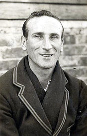A clean-shaven man with a thin face and wearing a blazer, and with a dark scarfe around his neck is smiling at the camera.
