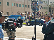 An African American male wearing a black suit and blue shirt is shown with both his hands crossed while staring at the ground. There is a Caucasian male wearing a cap and a light blue shirt standing behind a video camera that is placed on camera stand. The video camera is pointed at the African American male. In the background, there is a building, a blue car parked in front of the side walk in front of the building and a blue van can be seen on the side walk. There are also groups of people standing around the building.