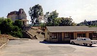 A brown one-story motel with a car parked outside. A big mansion is seen in the background.