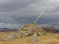A pile of stones in a grassy clearing in late autumn. A pole with a small windsock protrudes from its right