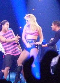 Image of a blond female performer. She is wearing a pink tank top and a pair of black boots. She is looking to her backup dancer at her right. He is wearing a pink shirt and black shorts.