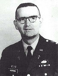 Head and shoulders of a middle-aged man with closely cropped hair and horn-rimmed glasses. He is wearing a dark military jacket with a parachute badge on the left breast and a Christian cross under a "U.S." pin on each lapel.