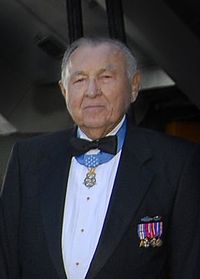 Head and shoulders of an elderly man with a round face, wearing a tuxedo with military medals pinned to the chest and one medal hanging from a light blue ribbon around his neck.