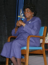 Claudette Colvin on a chair with a purple suit and glasses speaking into a microphone by a flag