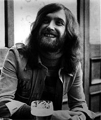A young, bearded man, wearing a jacket over a striped turtleneck sweater, sits a table presumably in a bar or pub.