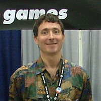 A man wearing multicolored shirt smiles at the camera. Behind him stands a blue and white background, above which is an excerpt of logo which reads "games".