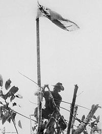 A kneeling man in military uniform raising a flag up a flag pole.