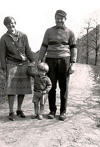 a man, wearing a striped sweater and trousers and a hat, with a woman, wearing a skirt and a cardigan, holding the hand of a boy wearing shorts, on a walking path