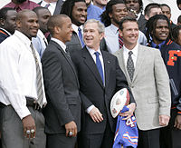 Photo depicts a smiling Chris Leak, a 22-year-old African-American man, together with his Florida Gators football teammates, coach Urban Meyer, and U.S. President George W. Bush, who is holding a Gators football jersey and commemorative football.