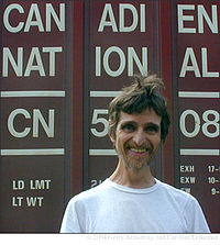 Head and shoulders photograph of a bearded smiling man in white T-shirt standing in front of a freight car bearing the words "Canadien National" and loading information