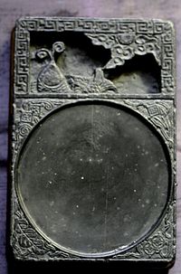 A black, rectangular stone tablet with two sections. The top quarter section contains a carving of a fish, and the bottom three fourths contain a shallow, circular indent for the ink.