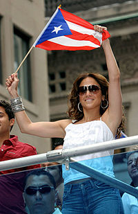 A middle aged, brown-haired woman, wearing brownish glasses, white T-shirt and blue jeans, waves the flag of Puerto Rico.