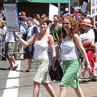 Photo of two women holding hands, one with short hair and the other with long, both wearing white spaghetti strap blouses that read "Bride", and green skirts while the one on the left holds a sign reading "Married March 11" with a blown-up reproduction of a marriage license. They are walking down a street with other people walking behind them holding signs as if for a parade or demonstration
