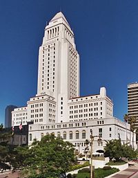 A tall, white, stone building set against a cloudless blue sky. The base is thicker than the top and is surrounded by smaller buildings of a similar material