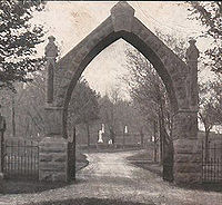 A slightly pinkish black and white photograph of a pointed stone entry arch with iron fencing on either side and a cemetery with grave monuments beyond
