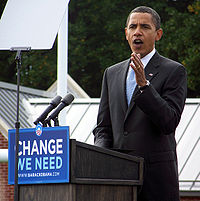 Full body photograph of a middle-aged man wearing a black suit and a blue tie speaking in front of a teleprompter. A sign spelling "Change we need" is seen in the podium at which the man is speaking.