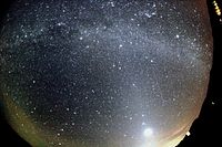 A meteor strikes the bottom left, while the Milky Way arcs overhead and a dawn-like light lines the lower horizon. The image was taken through a curved lens.