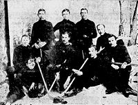 Men wearing hockey sweaters assembled in three rows in front of a painting of a forest scene. Several are holding hockey sticks