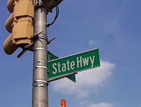 A green street sign reading State Highway attached to a traffic light pole.