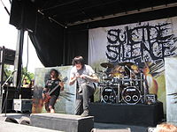 Two men are depicted on an outdoor stage set during the daytime, the man on the left plays bass while the man on the right is shown holding a microphone.