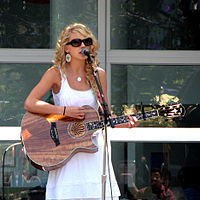 A blond female, clothed by a white sundress and large sunglasses, in a semi-right profile view playing a wooden acoustic guitar. Behind her appears a window in daylight.