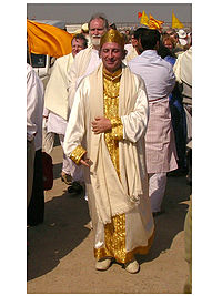 A photograph of a middle-aged caucasian male who dressed in white silk robes with gold trim, wearing a golden crown and standing outdoors in a crowd.