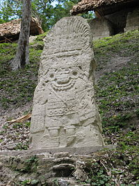 An upright stone shaft with the frontal image of a warrior with spear and shield and elaborate feathered headdress.