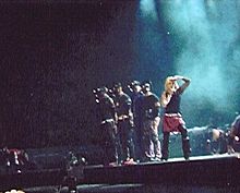A faraway image of a blond woman on stage. She wears red skilts, black top and her hair is short. On her right four male dancers stand looking to the front of the stage. The woman puts her left hand in front of her face, shedding her eyes. The backdrop is bluish in color.