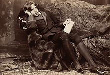 A tall man rests on a chaise longue, facing the camera. On his knees, which are held together, he holds a slim, richly bound book. He wears knee breeches which feature prominently in the photograph's foreground.