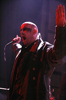 Head and upper body shot of a male singer, with a shaved head and blackened eyes, performing on stage. He is wearing a double-breasted military overcoat with a large collar.