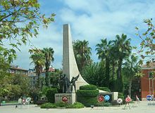 A tall sweeping stone triangle projects skyward behind the statues of a man and two children in bronze on a smaller podium. Around the base are place several wreathes with logos. Palm trees surround the scene.