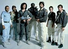 The seven principal cast members of the film stand in front of a white backdrop, in costume and holding prop weapons from the film.