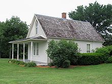 The exterior of the small home. Visible damage is seen near the front porch foundation, although restoration is also evident in metallic reinforcement of the shingled roof and chimney area.