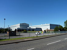 A photograph of a modern factory with two large industrial buildings standing side by side and a loading area at the front. The name of the company is written in red lettering at the front of each building. A colummn of steam rises in the background