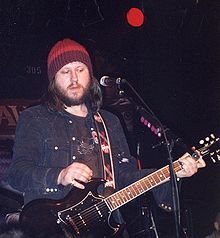 A man playing a guitar and singing on stage. He is wearing a denim jacket and woolen cap