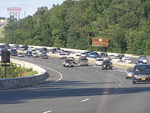 A tree lined six lane freeway with congestion on the right side of the road surrounded by trees. To the right, a brown sign reads Maryland Route 450 Bladensburg Annapolis with an arrow pointing to the upper right. A sign for the Capital Plaza Mall is in the distance