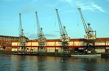 A long two storey building with 4 cranes in front on the quayside. Two tugboats are moored at the quay.