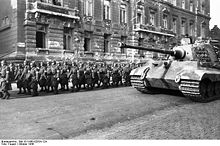 A large tank with sloped frontal armor and a flat faced turret, by a column of marching soldiers wearing overcoats and helmets, in a wide city street. A large building to the rear shows the scars of battle.