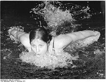 Woman emerging above the surface of the water in a swimming pool to take a breath as she is stroking butterfly. Her arms are behind he shoulders and moving forward for another stroke. she is capless and goggleless and has dark hair.