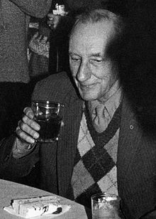Older man wearing a chequered sweater under a suit coat. He is sitting at a table, drinking, with a piece of cake on a napkin in front of him.