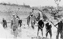 Six men plow the earth in a sinkhole while another walks carrying empty baskets. Three others are standing and walking in the background.