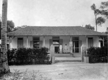 Two men stand on the porch of a single story building behind an open gate lined with bushes