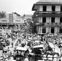 Crowd at a busy street intersection. There are horse-drawn carriages in the foreground while a three story building (with the sign "Kam Leng") and a single story building (with the sign "Chunghua Bioscoop") stand in the background on adjacent corners of the intersection.