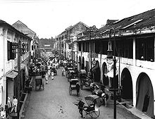 High angle view of a street lined with two and three story structures. The street is filled with crowds, including horse-drawn and hand-pulled carriages.