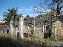 Worn brick walls and arches with a building visible to the left and vegetation in the foreground