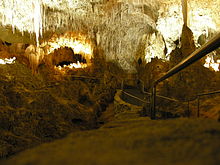 A picture of a partially illuminated underground cave with a jagged rock ceiling and a walking-bridge extended into the cavern.