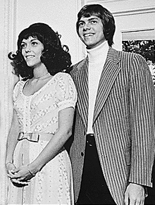 black and white picture of an attractive man and woman in their twenties; the woman on the left is clasping her hands and wearing patterned, short-sleeved dress with a bow, and the man on the right is wearing a striped jacket.