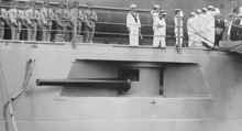 photograph of a cannon mounted in an armored bulge in the side of a ship, just below a deck with a number of sailors.