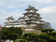 A white castle with a large five-storied main  tower and two smaller towers all built on a stone base.