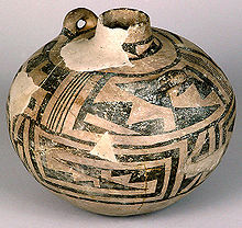 A nearly spherical ceramic vessel, painted with a network of black triangles and lines over a tan surface; many chips and cracks reveal a beige substratum. The top tapers to a short and narrow cylindrical neck. A toroidal carrying handle protrudes outward near it.